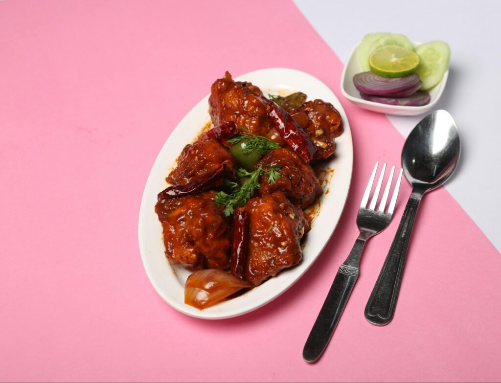 Delicious Chicken Manchurian garnished with herbs, served on a pink background with cutlery.